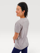 Woman wearing a light grey Iksplor merino wool t-shirt with short sleeves, side view against a white background. #color_trail dust