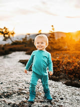 A smiling toddler wearing a teal Iksplor merino wool zip romper and boots, standing on a rocky path at sunset.