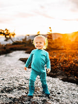 Toddler wearing a bright teal Iksplor merino wool zip romper while standing on a rocky path at sunset. #color_tidepool