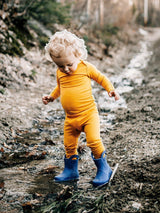 Toddler wearing a mustard yellow Iksplor merino wool bodysuit and blue rain boots playing in a muddy outdoor path. #color_yellowstone