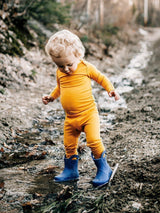 Toddler wearing a mustard yellow Iksplor merino wool bodysuit and blue rain boots playing in a muddy outdoor path. #color_yellowstone