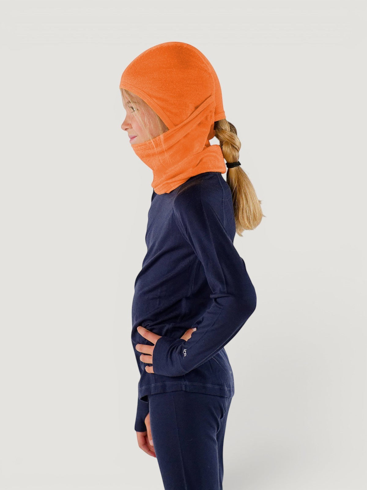 Child wearing an orange Iksplor merino wool balaclava with a ponytail hole, side view. #color_poppy