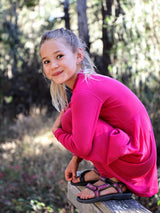 A young girl wearing a bright pink Iksplor merino wool adventure dress, crouching outdoors in a forest setting. #color_peak pink