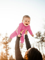 Iksplor baby wearing a bright pink merino wool long sleeve onesie being lifted outdoors in the snow . #color_alpenglow