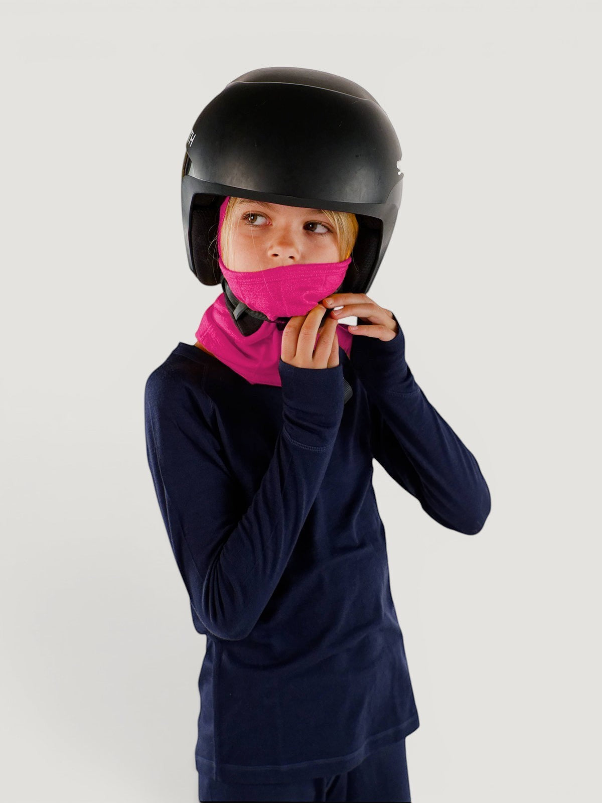 Child wearing a bright pink Iksplor merino wool balaclava with a helmet and navy long-sleeve base layer #color_peak pink