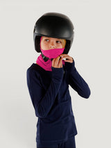 Child wearing a bright pink Iksplor merino wool balaclava with a helmet and navy long-sleeve base layer #color_peak pink