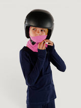 Child wearing a pink Iksplor merino wool balaclava with a ponytail hole under a black ski helmet. #color_alpenglow