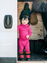 Toddler wearing a bright pink Iksplor merino wool bodysuit and matching pants standing in a camper van doorway. #color_peak pink