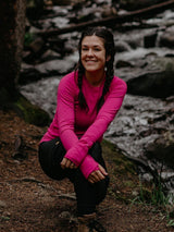 Woman wearing a bright pink Iksplor merino wool long sleeve top with thumbholes while crouching by a forest stream. #color_peak pink