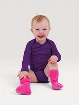 Happy baby wearing a long-sleeve purple Iksplor merino wool bodysuit and bright pink socks #color_huck-it-berry