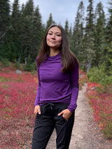Woman wearing a purple Iksplor merino wool long sleeve top on a forest trail with red autumn foliage.  #color_huck-it-berry