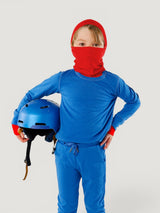 Child wearing a red Iksplor merino wool balaclava with a ponytail hole and a blue helmet in hand #color_campfire
