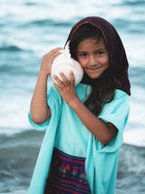 Young girl wearing an Iksplor x SURreal turquoise terry cloth towel poncho with a dark purple hood at the beach. #color_high tide