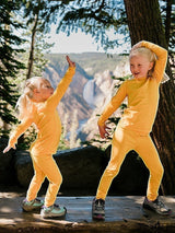 Two children wearing bright yellow Iksplor merino wool base layer leggings and tops dancing in a forest setting. #color_yellowstone